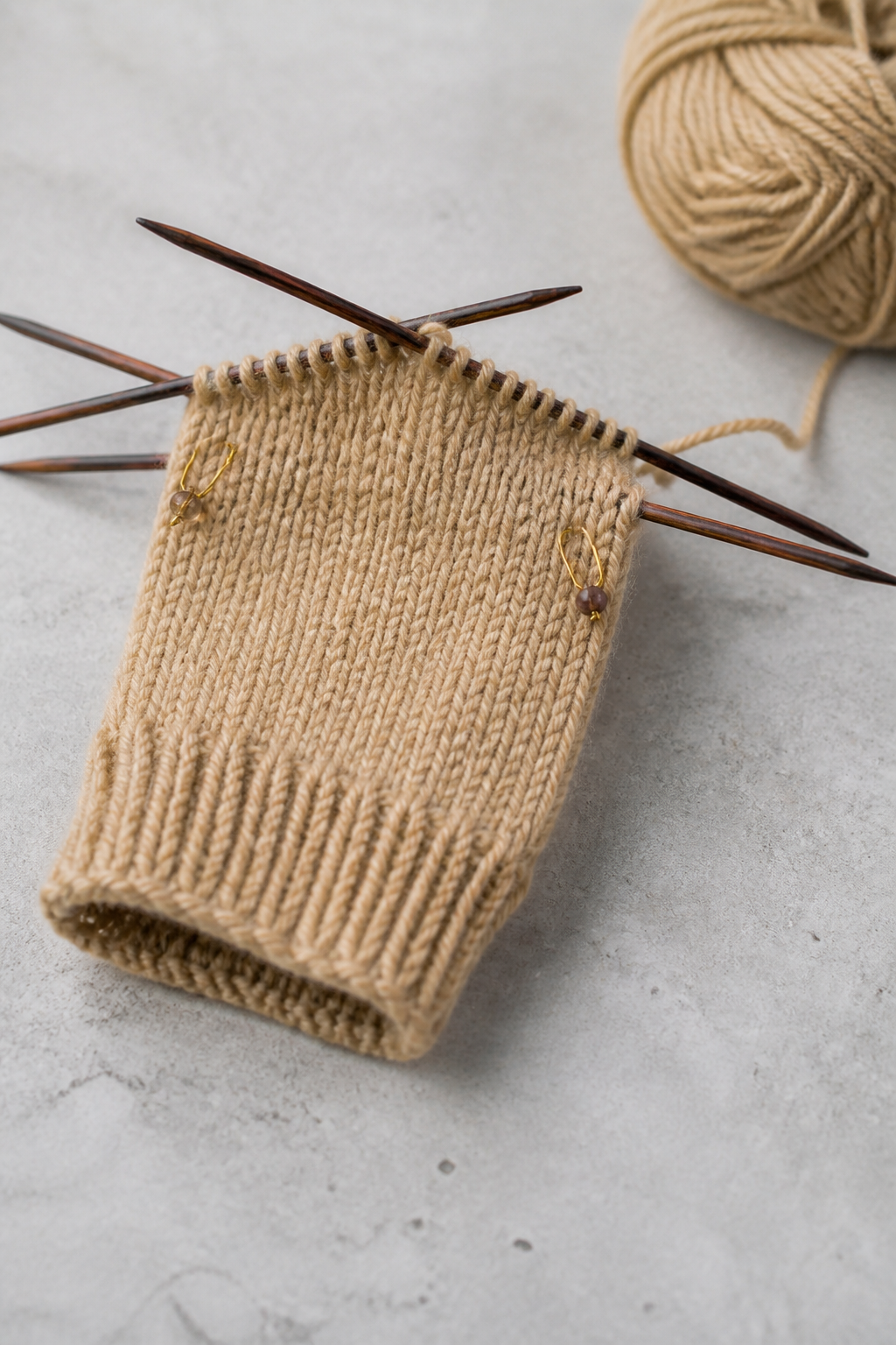 A part-knitted mitten on four double-pointed needles, with the thumb gusset increases marked by two stitch markers.